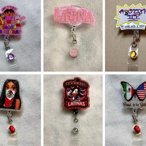 May include: Six retractable badge reels featuring various designs: a Dora the Explorer graphic with text, a pink glittery 'LATINA' sign, a yellow 'ABOLISH ICE' sign, a person wearing a Mexican flag mask, a Shadow character graphic, and a butterfly with flags.