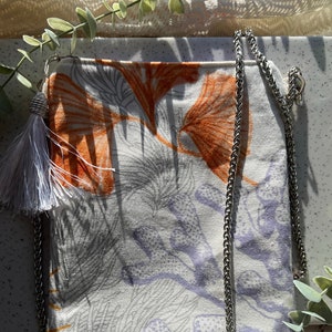 Puede incluir: Un bolso bandolera rectangular con una cadena plateada. El bolso presenta un fondo blanco con un estampado floral en naranja, gris y morado. Una borla gris cuelga de la esquina superior izquierda.