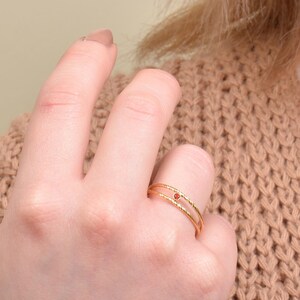 May include: A gold ring with a small red gemstone in the center. The ring is double-banded and has a delicate, textured design.