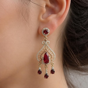 Peut inclure: Une boucle d'oreille chandelier dorée avec une pierre rouge ronde en haut, entourée de petits cristaux clairs. La goutte centrale est une pierre rouge en forme de poire, accentuée par trois pierres rouges plus petites en forme de poire.