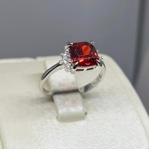 May include: A silver ring with a large, red, square-shaped gemstone surrounded by smaller, clear gemstones.
