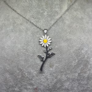 Puede incluir: Un collar de cadena de plata con un delicado colgante de margarita. La margarita tiene un centro blanco y pétalos amarillos.