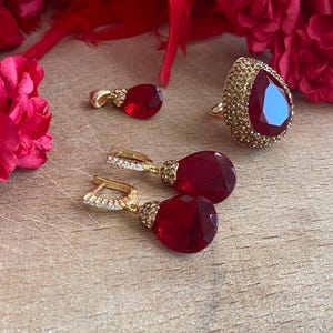 May include: A set of red gemstone jewellery featuring a ring, a pendant, and a pair of earrings. The jewellery is made of gold and features a teardrop-shaped red gemstone.