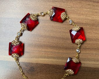 Pode incluir: Uma pulseira dourada com grandes pedras preciosas vermelhas retangulares facetadas. A pulseira é conectada por delicadas correntes douradas e acentuada com cristais transparentes menores e detalhes em filigrana dourada. O fecho também é dourado.
