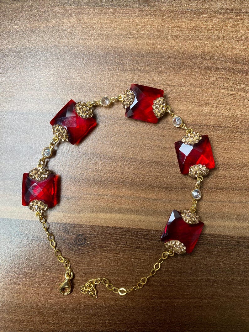 Pode incluir: Uma pulseira dourada com grandes pedras preciosas vermelhas retangulares facetadas. A pulseira &eacute; conectada por delicadas correntes douradas e acentuada com cristais transparentes menores e filigrana dourada. O fecho tamb&eacute;m &eacute; dourado.