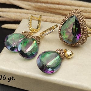 Puede incluir: Un conjunto de joyas con un anillo, pendientes y un colgante con piedras preciosas iridiscentes en forma de lágrima. El anillo y los pendientes tienen engastes dorados con pequeñas piedras transparentes. El colgante también tiene un engaste dorado. El conjunto se exhibe en una caja blanca.