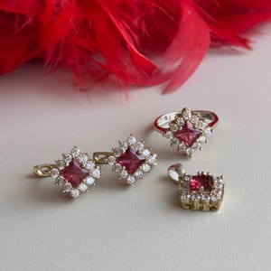 May include: A set of jewelry featuring a ring, earrings, and a pendant. Each piece has a square-shaped red gemstone surrounded by smaller clear gemstones.