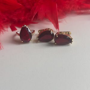 Puede incluir: Un anillo dorado y dos pendientes a juego, cada uno con una gran piedra preciosa roja oscura en forma de lágrima. Las joyas están sobre una superficie blanca con un fondo de plumas rojas.