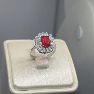 May include: A silver ring featuring a large, rectangular, vibrant red gemstone at the center, surrounded by a halo of smaller, rectangular, clear stones. The ring is displayed on a cream-colored surface.