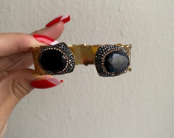 Pode incluir: Um bracelete de punho dourado com duas pedras ovais pretas engastadas em um leito de strass brilhantes.