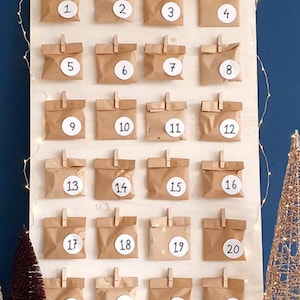May include: A wooden board with 24 brown paper bags, each with a white circle containing a number from 1 to 24. The board is decorated with white fairy lights.