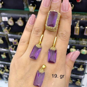 Puede incluir: Un anillo de oro con una gran piedra preciosa púrpura rodeada de piedras preciosas transparentes más pequeñas. El anillo se combina con un par de pendientes a juego y un colgante, todos con el mismo diseño. La joyería pesa 19 gramos.