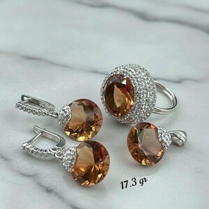 Puede incluir: Un conjunto de joyas con un anillo, un colgante y pendientes. El anillo y los pendientes tienen un engaste plateado con una piedra grande, ovalada y de color ámbar. El anillo está rodeado de pequeñas piedras transparentes. El colgante tiene un diseño similar.