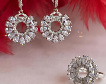 Könnte beinhalten: Ein Paar silberfarbene Ohrhänger und ein passender Ring, jeweils mit einem kreisförmigen Design, das mit klaren, birnenförmigen Steinen in einem Strahlenmuster besetzt ist. Die Ohrringe haben Klappbügel mit kleinen, pavé-gefassten Steinen.