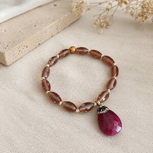 May include: A beaded bracelet featuring translucent, faceted, oval-shaped brown beads and a single metallic gold bead. A large, teardrop-shaped, deep magenta pendant with a dark, textured cap hangs from the bracelet, adding a bold focal point.