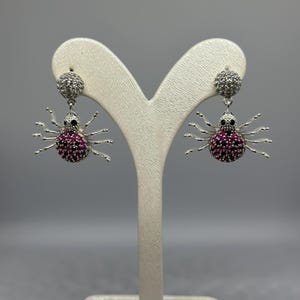 May include: A pair of silver spider earrings with pink and white cubic zirconia stones.