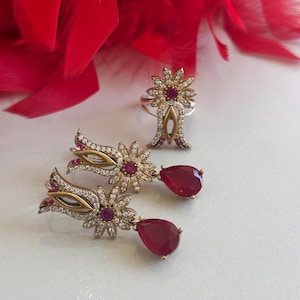 Puede incluir: Un conjunto de pendientes y un anillo de plata con detalles de piedras preciosas rojas. Los pendientes presentan un diseño floral con una piedra preciosa roja colgante. El anillo tiene un diseño floral similar con una piedra preciosa roja en el centro.