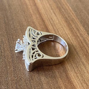 May include: A silver ring with a large, clear stone in the center and intricate filigree detailing around the stone. The ring is set on a wooden surface.