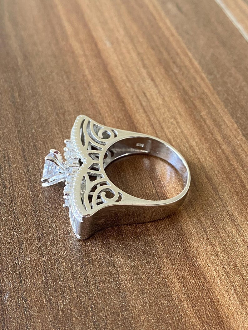 May include: A silver ring with a large, clear stone in the center and intricate filigree detailing around the stone. The ring is set on a wooden surface.