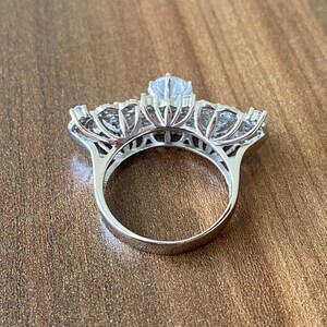 May include: A silver ring with a large, clear center stone and smaller stones surrounding it. The ring is set in a delicate, floral design.
