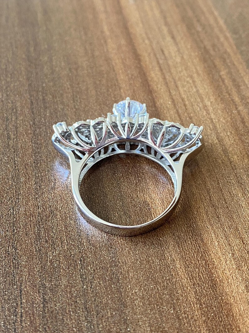 May include: A silver ring with a large, clear center stone and smaller stones surrounding it. The ring is set in a delicate, floral design.