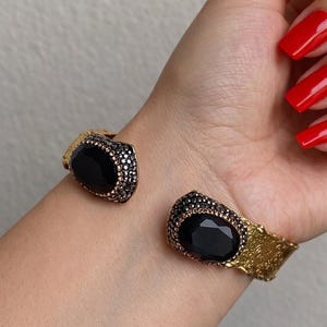 Può includere: Bracciale a polsino dorato con due grandi pietre nere ovali. Le pietre sono circondate da pietre più piccole e scure. Il bracciale ha un design strutturato.