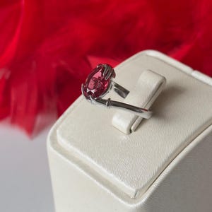 May include: A silver ring with a large, oval-cut, deep pink gemstone. The gemstone is set in a prong setting. The ring is displayed on a white, textured surface against a blurred red background.