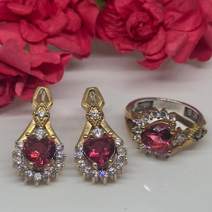 May include: A set of jewellery featuring a heart-shaped red gemstone surrounded by clear stones. The set includes a ring and a pair of earrings.