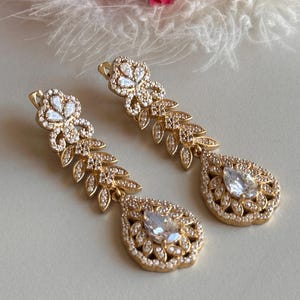 May include: A pair of gold dangle earrings with clear crystal stones. The earrings feature a teardrop-shaped design with a floral motif.