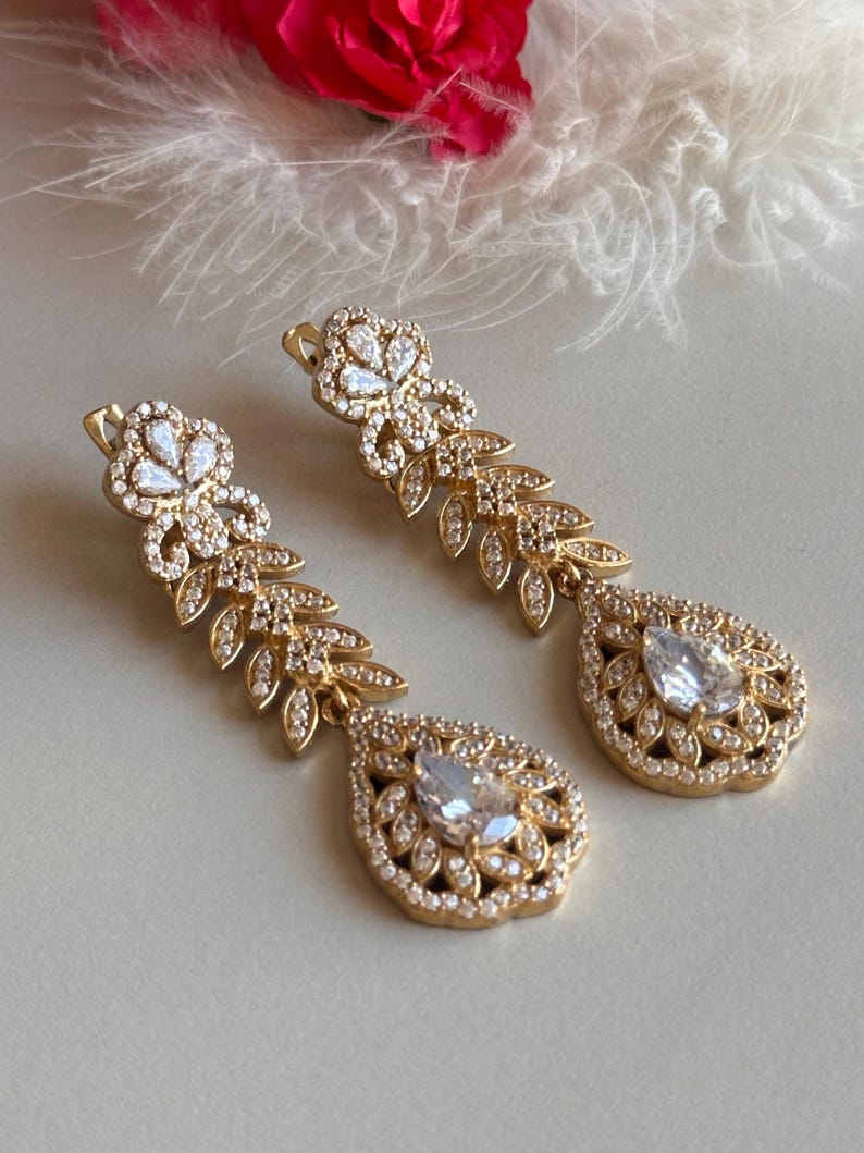 May include: A pair of gold dangle earrings with clear crystal stones. The earrings feature a teardrop-shaped design with a floral motif.