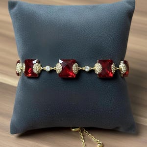 Könnte beinhalten: Ein goldfarbenes Armband mit großen, quadratischen, rubinroten Edelsteinen. Das Armband ist mit kleineren, klaren Kristallen und goldfarbenen Verzierungen versehen. Die verstellbare Kette ist ebenfalls goldfarben. Das Armband wird auf einem dunkelgrauen Kissen präsentiert.