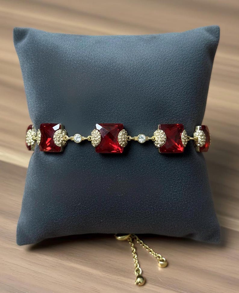 Pode incluir: Uma pulseira dourada com grandes pedras preciosas quadradas de cor vermelho rubi. A pulseira &eacute; adornada com cristais transparentes menores e enfeites dourados. A corrente ajust&aacute;vel tamb&eacute;m &eacute; dourada. A pulseira &eacute; exibida em uma almofada cinza escuro.