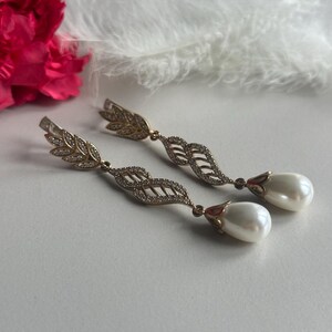 May include: A pair of gold tone dangle earrings with a pearl drop. The earrings feature a delicate leaf design with clear stones.