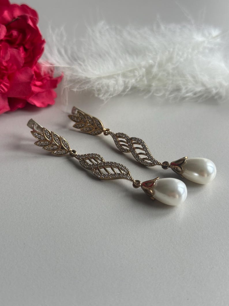 May include: A pair of gold tone dangle earrings with a pearl drop. The earrings feature a delicate leaf design with clear stones.