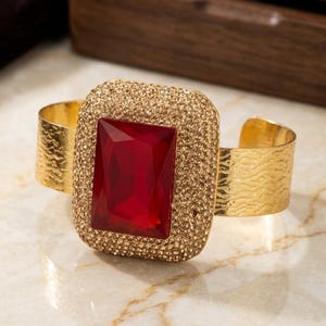 May include: A gold-tone cuff bracelet featuring a large, rectangular, faceted red stone at the center. The stone is surrounded by a wide, textured border encrusted with numerous small, shimmering gold-tone crystals. The band has a wavy texture.