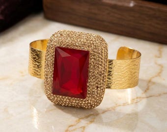 Könnte beinhalten: Ein goldfarbenes Armband mit einem großen, rechteckigen, facettierten roten Stein in der Mitte. Der Stein ist von einem breiten, strukturierten Rand umgeben, der mit zahlreichen kleinen, schimmernden goldfarbenen Kristallen besetzt ist.