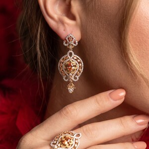 May include: A gold-toned drop earring and a matching ring, both featuring an oval-shaped orange center stone surrounded by intricate filigree metalwork and small clear pavé-set stones in a decorative, symmetrical, and ornate pattern.