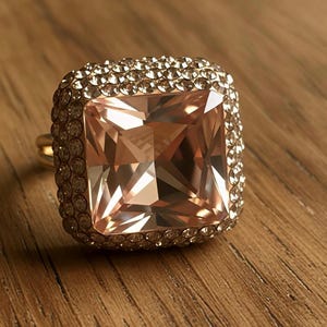 May include: A large, square-cut peach-colored gemstone ring with a halo of smaller, clear stones set in a gold band. The ring is displayed on a wooden surface, showcasing its facets and sparkle.