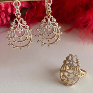 May include: A pair of gold-tone drop earrings and a matching ring, each featuring an intricate filigree teardrop design adorned with small, clear, and yellow stones arranged in concentric circular patterns throughout the metal frame.