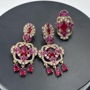 May include: A set of gold earrings and a ring with red and white stones. The earrings have a dangling design with three teardrop-shaped stones.