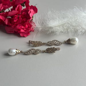 May include: A pair of gold earrings with a pearl drop and a leaf design. The earrings are on a white surface with a pink flower and a white feather.