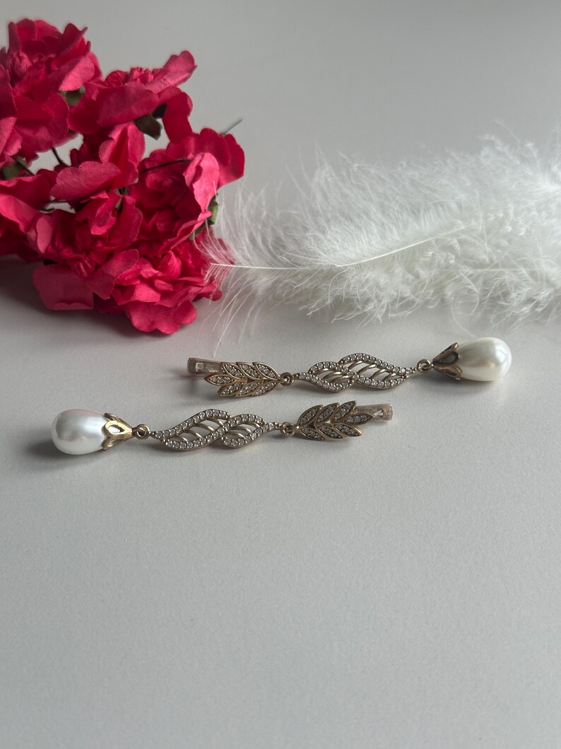 May include: A pair of gold earrings with a pearl drop and a leaf design. The earrings are on a white surface with a pink flower and a white feather.