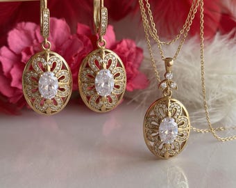 Könnte beinhalten: Ein goldfarbenes Schmuckset bestehend aus zwei ovalen Ohrhängern und einer passenden Halskette mit Anhänger. Jedes Teil zeigt ein filigranes Blumenmuster, das mit kleinen klaren Steinen und einem großen zentralen ovalen Stein besetzt ist.