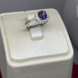 May include: A silver ring with a purple gemstone surrounded by a halo of smaller clear gemstones.
