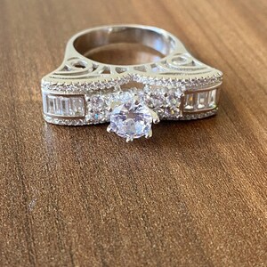 May include: A silver ring with a large, clear, round center stone and smaller, clear, rectangular stones on the band. The ring has a delicate, intricate design.