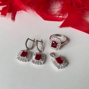 May include: A set of silver jewelry with red gemstone accents. The set includes a ring, a pendant, and a pair of earrings. Each piece features a square-shaped red gemstone surrounded by smaller clear gemstones.