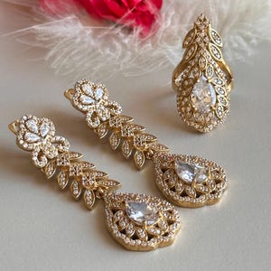 May include: A set of gold-tone earrings and a ring with clear cubic zirconia stones. The earrings are long and dangle, with a teardrop-shaped design. The ring is also teardrop-shaped and features a delicate leaf pattern.