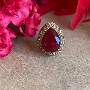 Könnte beinhalten: Ein großer, tropfenförmiger Ring mit einem tiefroten Edelstein in der Mitte. Der Ring ist von einem Kranz aus kleinen, goldfarbenen Kristallen umgeben. Im Hintergrund befinden sich rosa Blüten und eine Holzoberfläche.