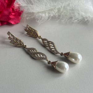 May include: A pair of gold earrings with a leaf design and a pearl drop. The earrings are on a white background.