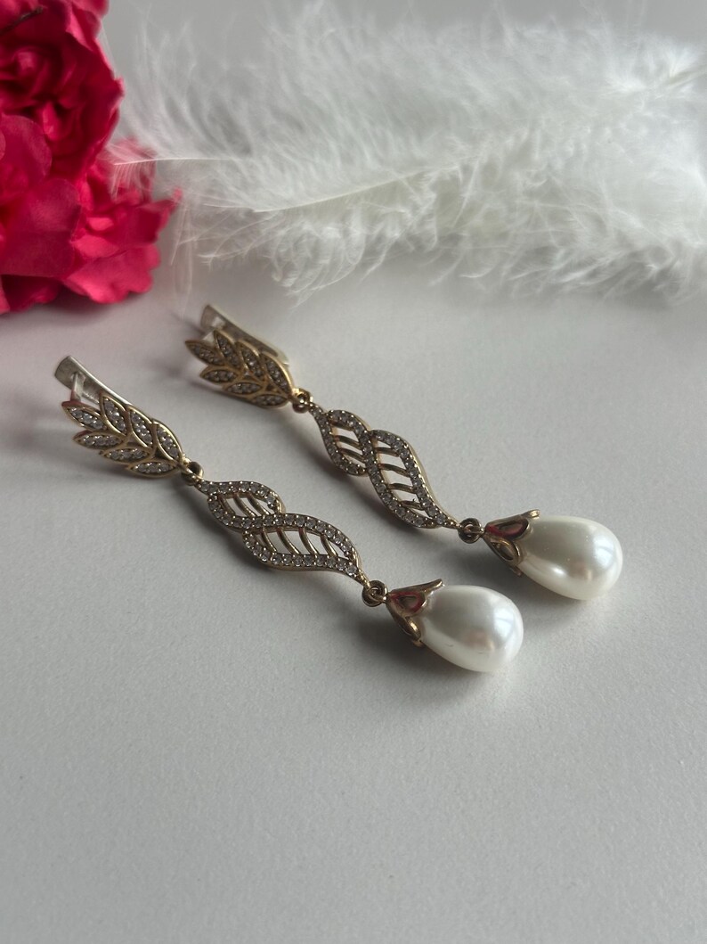 May include: A pair of gold earrings with a leaf design and a pearl drop. The earrings are on a white background.